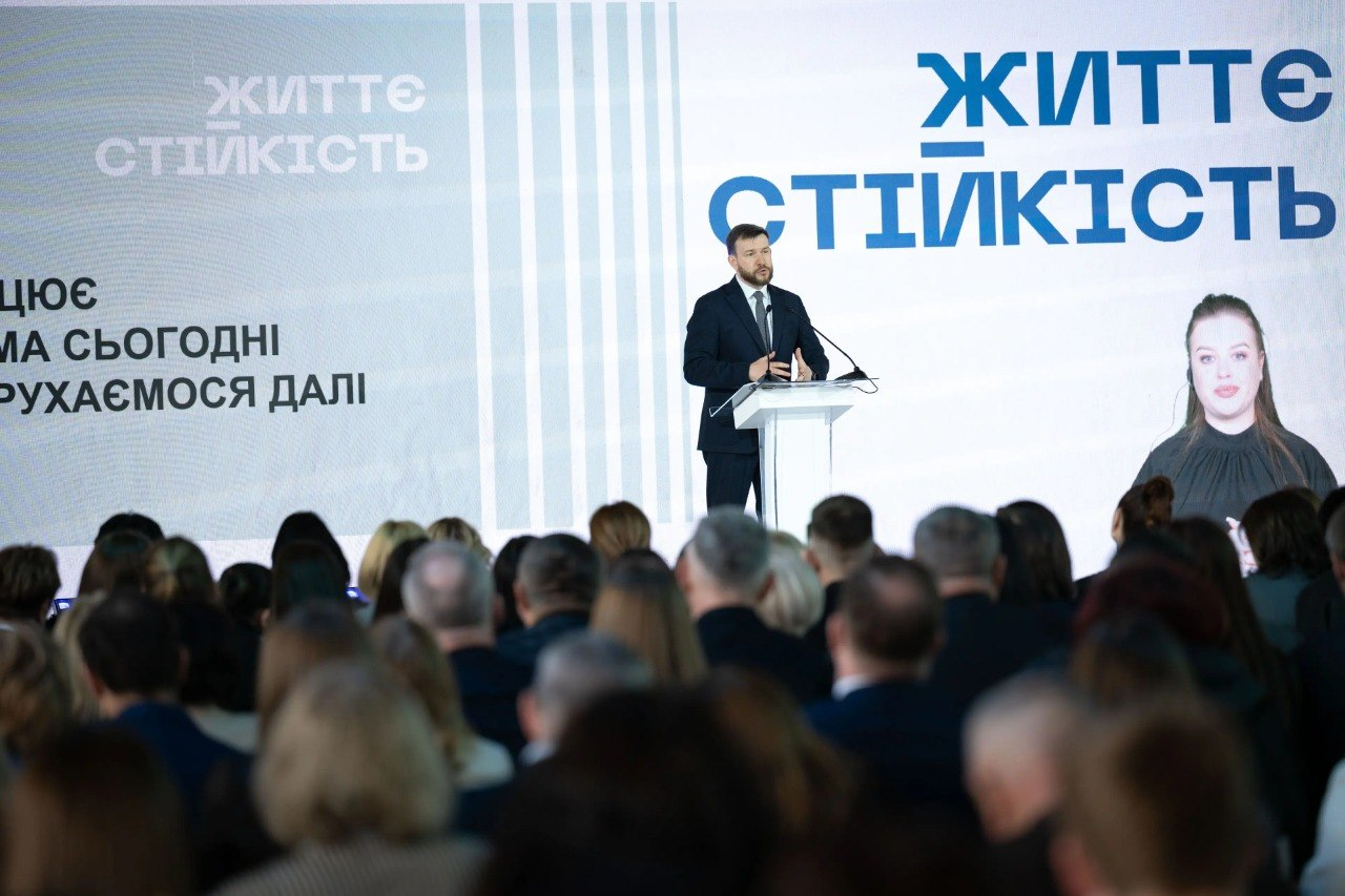 Виступ спікера за трибуною під час офіційного заходу перед великою аудиторією. На задньому плані — великий екран із написом «ЖИТТЄСТІЙКІСТЬ» та вікном із перекладачем жестовою мовою.