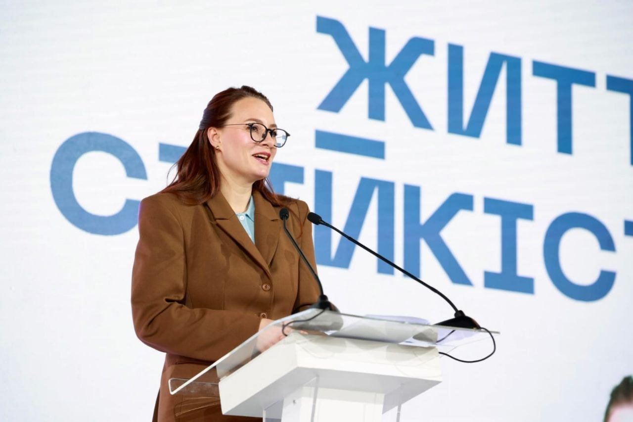 Жінка в коричневому костюмі та окулярах виступає за білою трибуною з мікрофонами. На задньому плані — великий світлий екран із частиною напису блакитного кольору «ЖИТТЄСТІЙКІСТЬ».