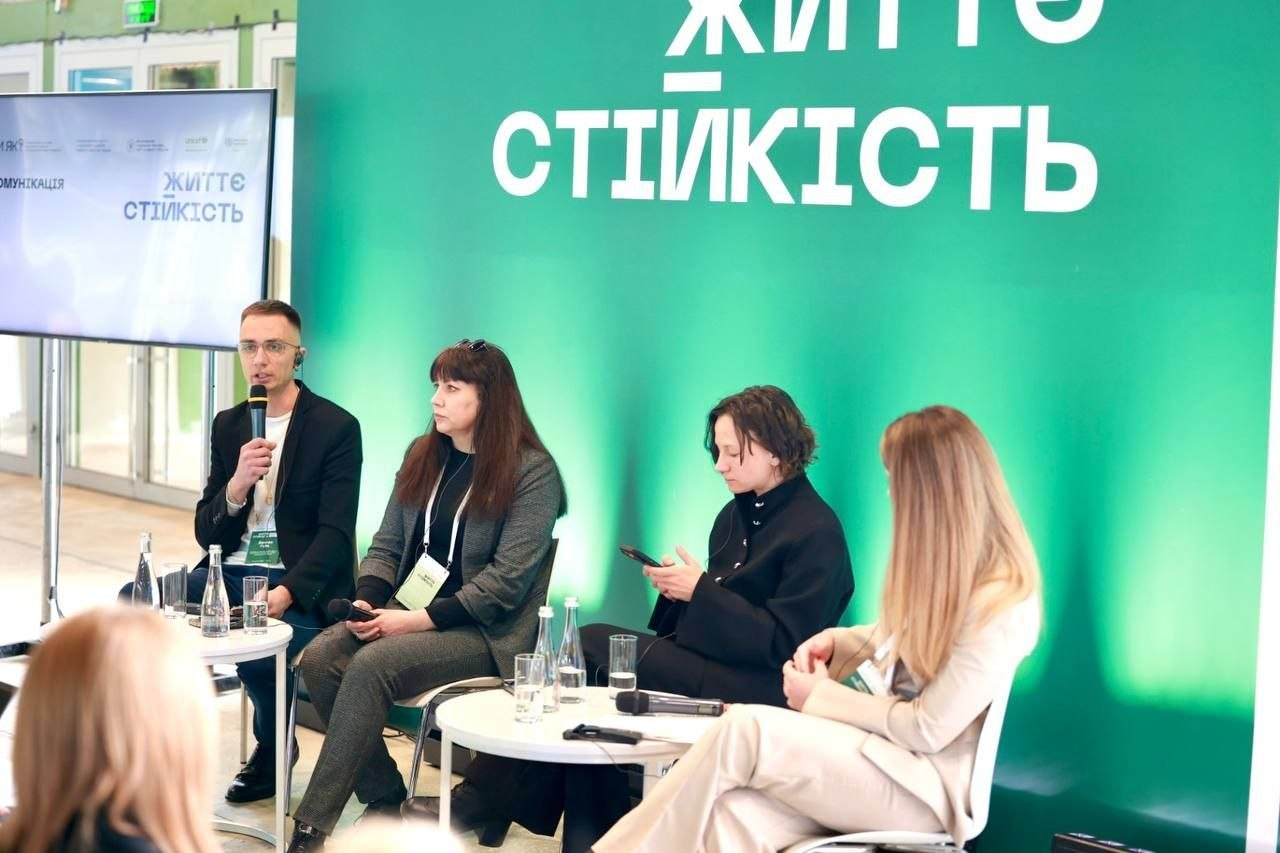Панельна дискусія за участю чотирьох осіб (чоловіка та трьох жінок) у форматі «запитання-відповідь». Учасники сидять у кріслах перед низькими круглими столиками на фоні яскраво-зеленого банера з написом «ЖИТТЄСТІЙКІСТЬ». Чоловік ліворуч говорить у мікрофон, поруч розташований монітор із презентацією.