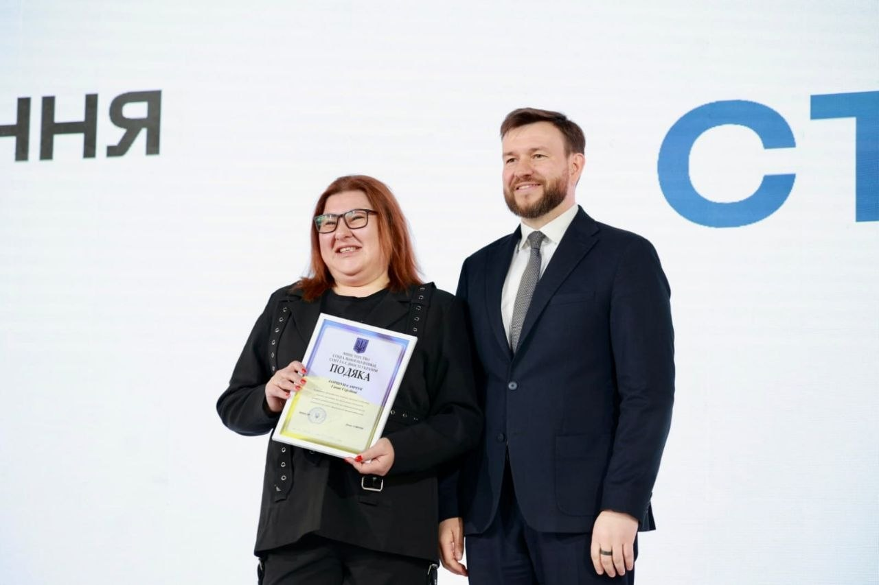 Урочистий момент нагородження: чоловік у костюмі вручає жінці почесну грамоту з написом «ПОДЯКА». Вони обоє усміхаються, позуючи для фото на фоні світлого банера з великими літерами.