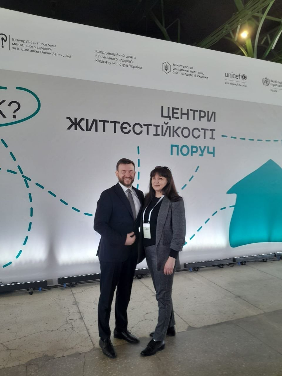 Чоловік та жінка в діловому вбранні позують на фоні великого білого банера з написом «ЦЕНТРИ ЖИТТЄСТІЙКОСТІ — ПОРУЧ». У верхній частині банера розміщені логотипи Всеукраїнської програми ментального здоров'я, Міністерства соціальної політики, ЮНІСЕФ та інших організацій.