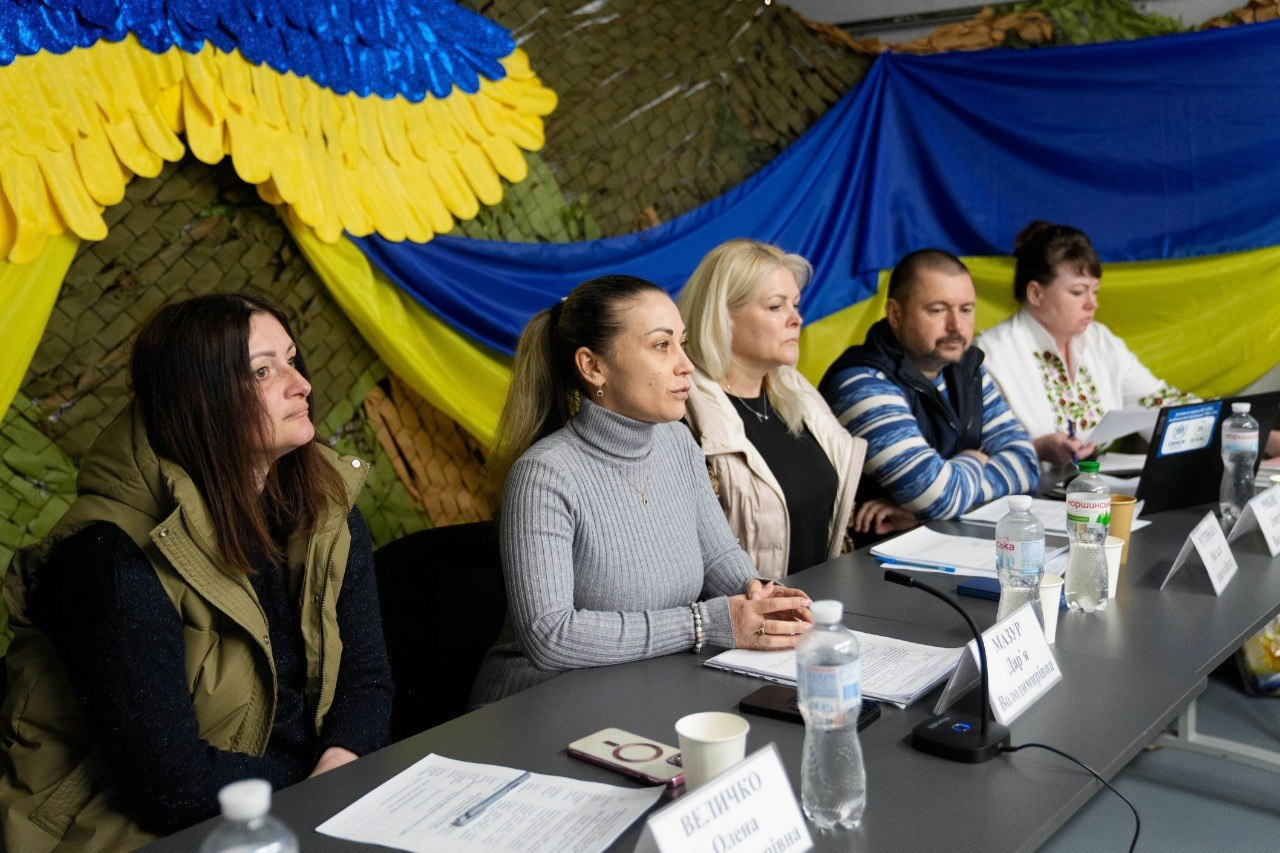 П'ятеро людей сидять за довгим столом під час офіційного заходу або пресконференції. Перед учасниками стоять таблички з іменами, пляшки з водою та документи. На задньому плані стіна завішена маскувальною сіткою та прикрашена великим жовто-блакитним ангелом і державним прапором України.