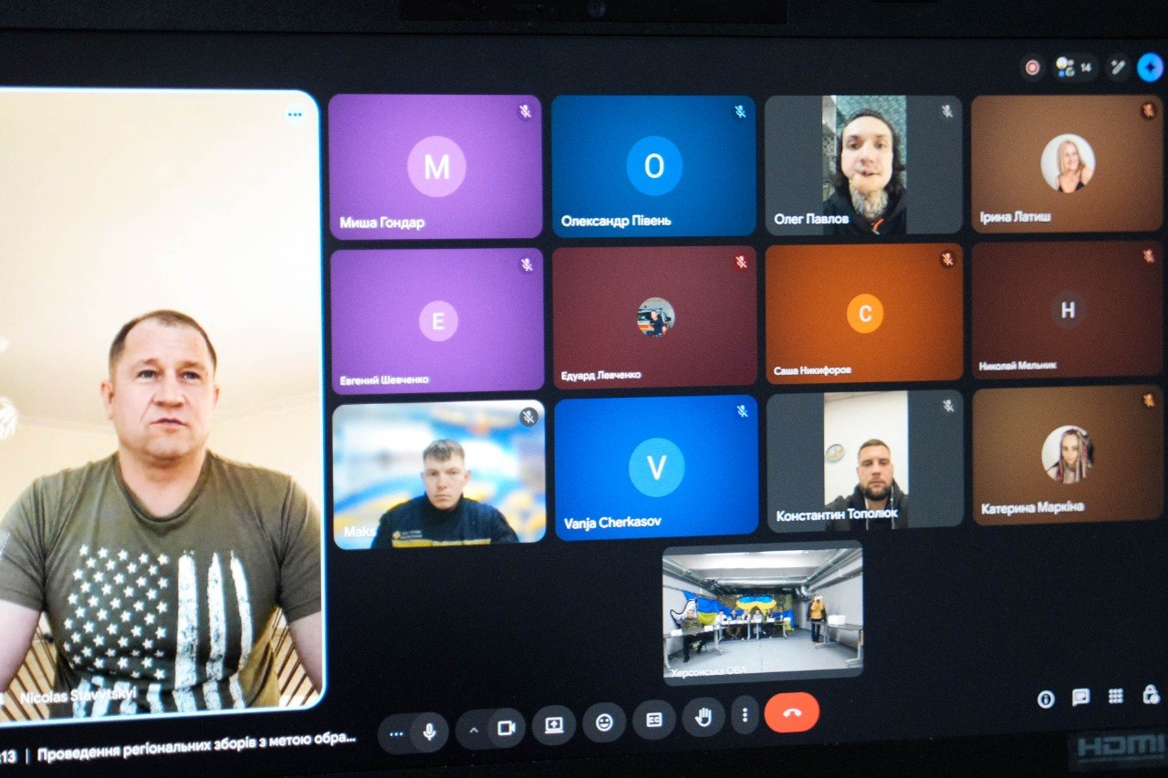 Скриншот вікна онлайн-конференції в Google Meet. Ліворуч у великому вікні зображений чоловік у футболці з патріотичним принтом. Праворуч — сітка з іншими учасниками зустрічі, дехто з яких увімкнув камеру, а інші представлені іконками з ініціалами. Внизу екрана видно рядок із назвою події про проведення регіональних зборів.