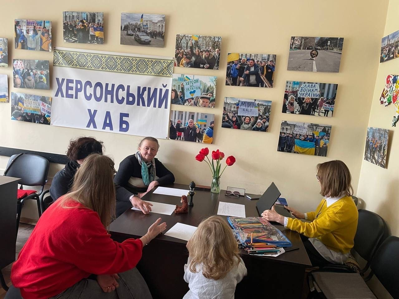 Жінки та дитина за робочим столом у приміщенні «Херсонського хабу». Стіна прикрашена патріотичними фотографіями та плакатами, на столі стоять червоні тюльпани.