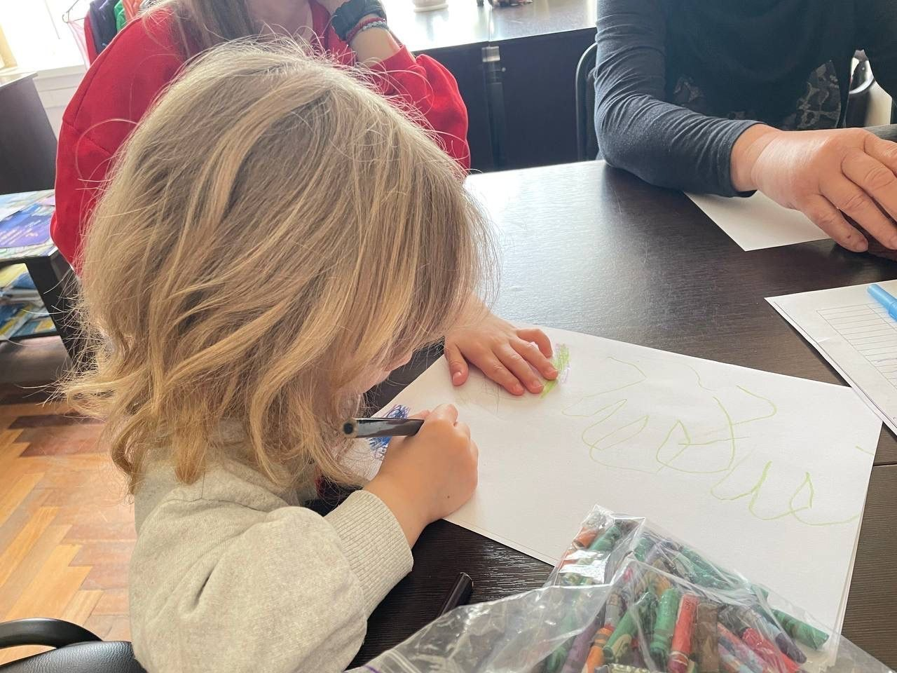 Дитина малює на папері за столом, на якому також лежить великий пакет із кольоровими восковими олівцями. Поруч сидять дорослі.