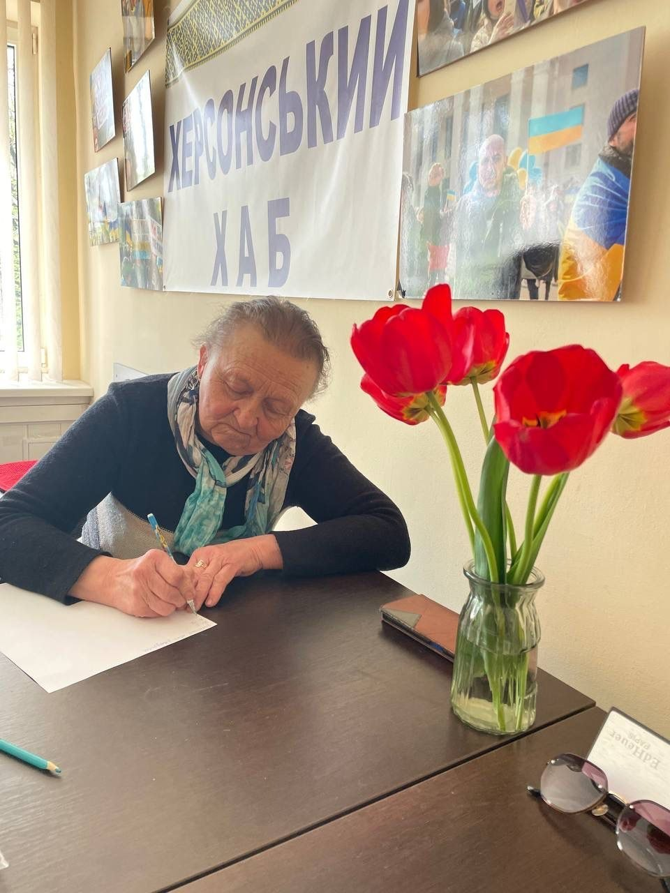 Жінка заповнює документи за столом, на якому стоїть ваза з червоними тюльпанами. На задньому плані — банер «Херсонський хаб» та патріотичні світлини на стіні.