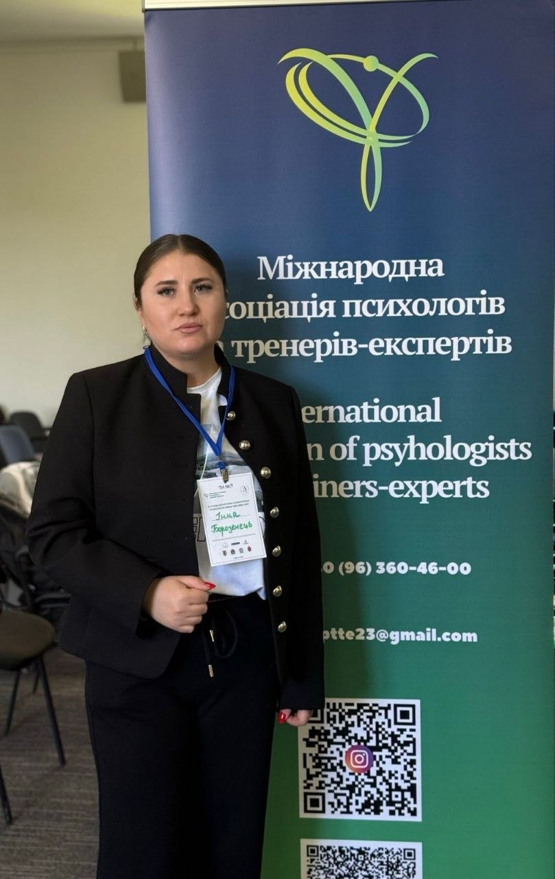 Жінка в діловому костюмі стоїть біля банера «Міжнародної асоціації психологів та тренерів-експертів». На банері вказано назву організації, контакти та QR-код.