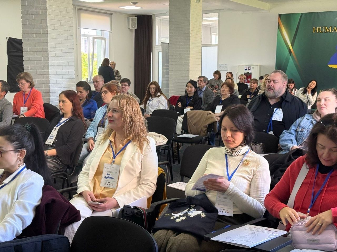 Група людей сидить у конференц-залі під час заходу. Учасники з бейджами на шиї слухають презентацію, дехто тримає в руках роздаткові матеріали.