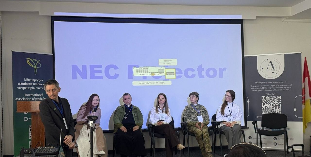 Група спікерів, серед яких є представники духовенства та військовий, сидить на сцені під час панельної дискусії. Позаду них — великий екран із написом «NEC Projector», а з боків розташовані інформаційні банери громадських організацій.