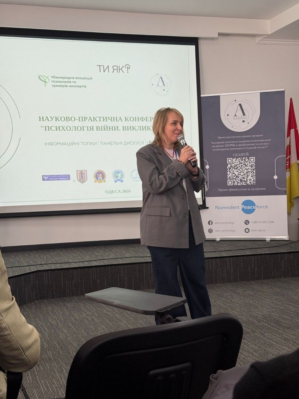 Спікерка виступає з мікрофоном на фоні екрана з презентацією науково-практичної конференції «Психологія війни. Виклики». Поруч стоїть інформаційний банер з QR-кодом та логотипами організацій-партнерів.