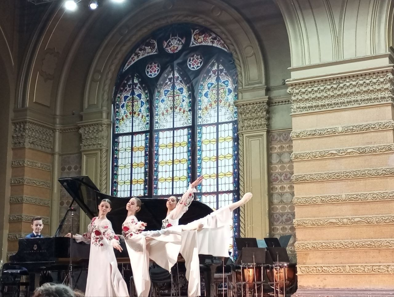 Виступ трьох танцівниць у білих сукнях з українською вишивкою на сцені з класичним архітектурним оздобленням. На задньому плані — юний піаніст за великим чорним роялем, масивне вітражне вікно та елементи оркестру.