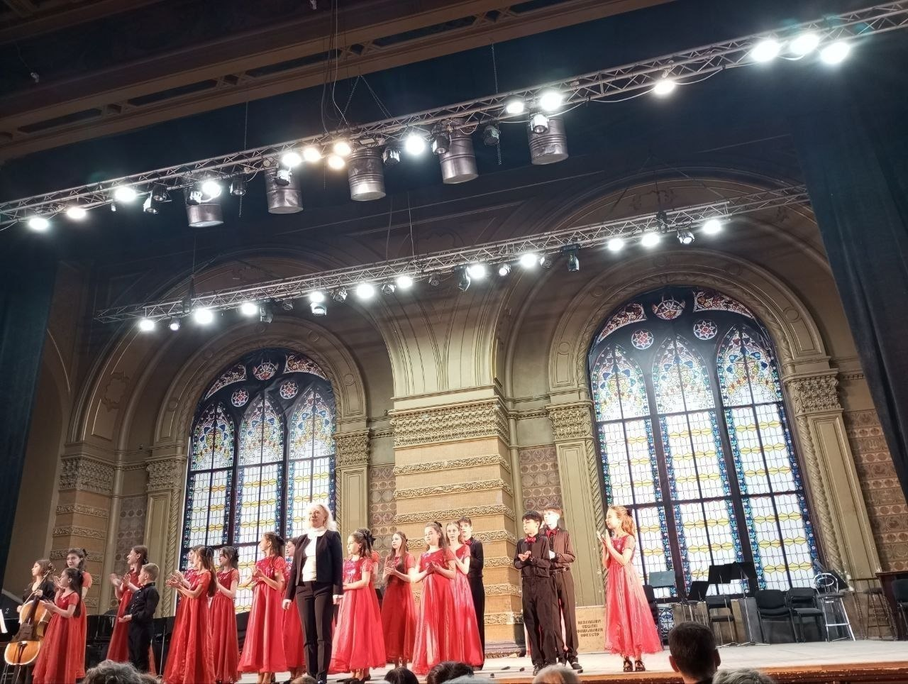 Виступ дитячого хору та юних музикантів на великій сцені. Учасники в ошатному вбранні — дівчата у довгих червоних сукнях та хлопці у темних костюмах — стоять під яскравим світлом софітів. На задньому плані видно величні вітражні вікна та класичне архітектурне оздоблення зали.