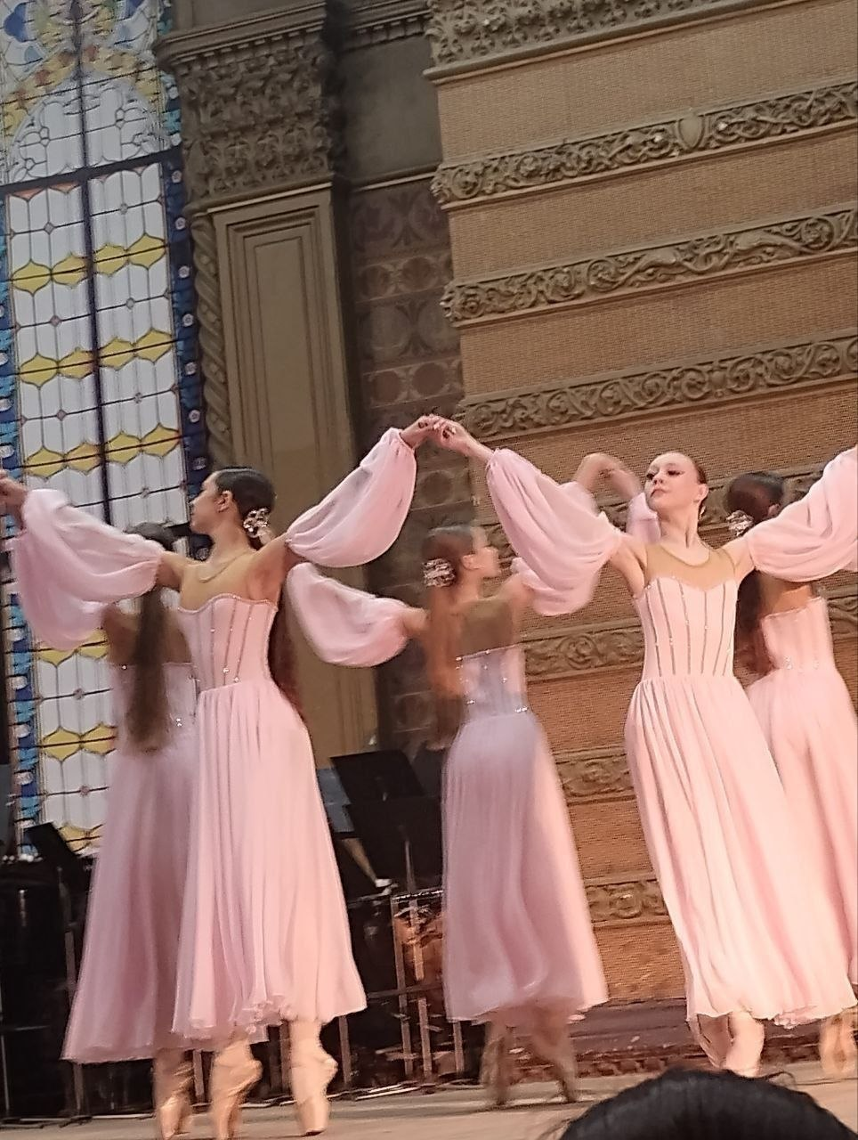 Груповий виступ балерин на сцені в інтер’єрі з класичною архітектурою. Танцівниці у ніжно-рожевих сукнях із пишними рукавами та пуантах виконують хореографічний номер на фоні великого вітражного вікна та оздоблених колон.