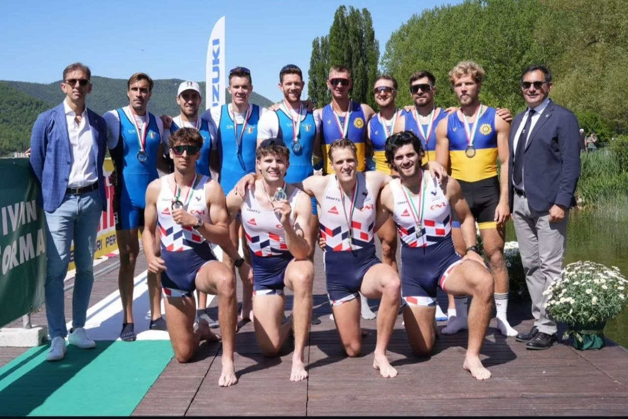 Групове фото спортсменів-веслувальників та офіційних осіб на дерев’яному причалі біля водойми. Атлети у спортивній формі різних команд з медалями на грудях позують на фоні мальовничого пейзажу з горами та деревами.