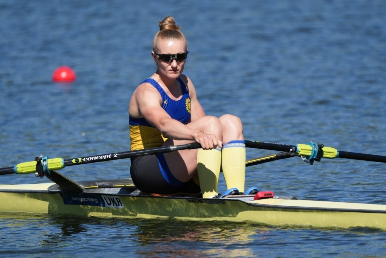 Спортсменка у синьо-жовтій формі та сонцезахисних окулярах веслує в одиночному човні на відкритій воді. На фоні водної гладі помітно червоний сигнальний буй, а на борту човна — маркування «UKR».