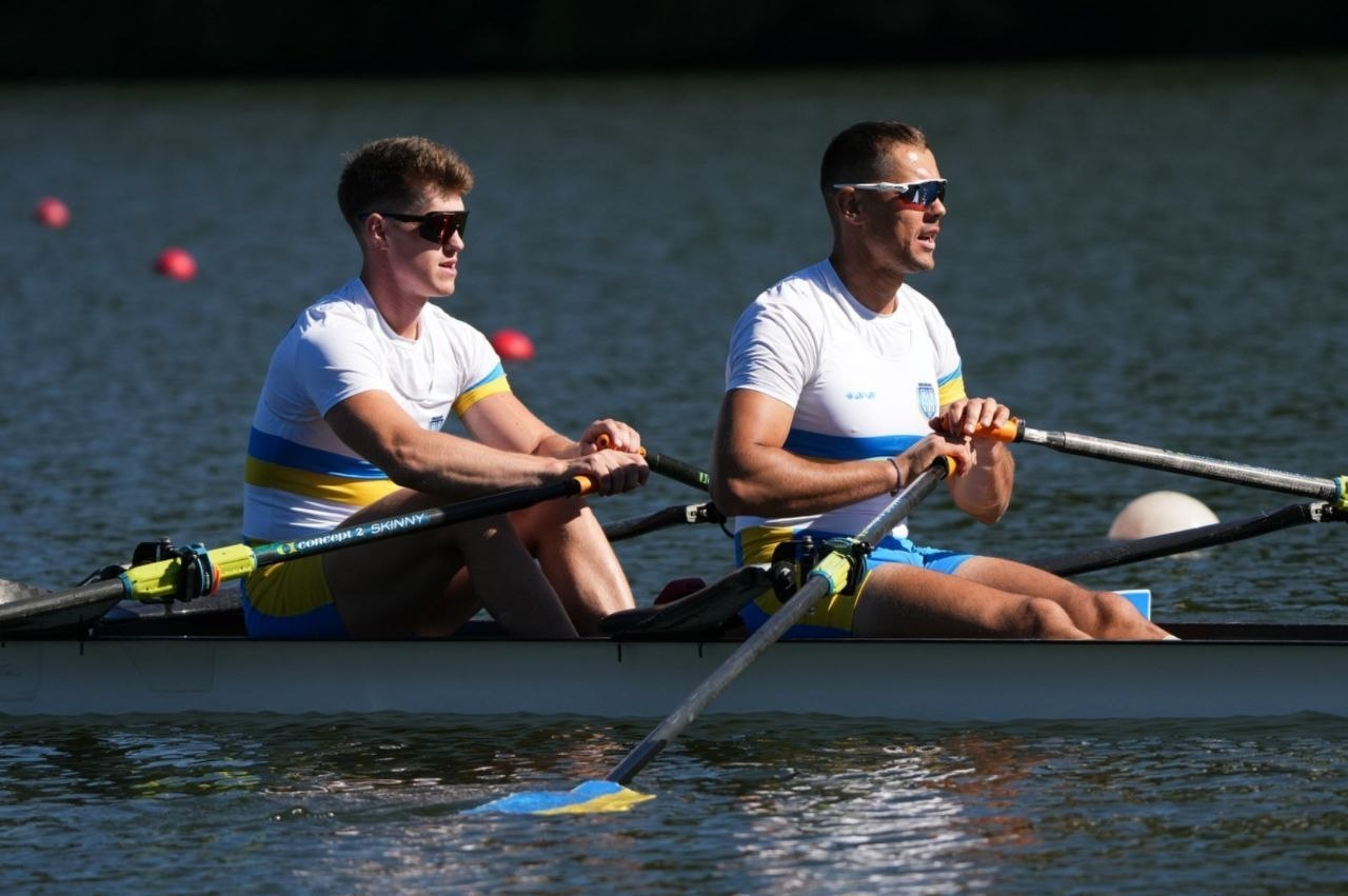 Двоє спортсменів у біло-синьо-жовтій формі та сонцезахисних окулярах синхронно веслують у човні-двійці на відкритій водоймі. На задньому плані видніються червоні та білі буї, що позначають водну трасу.
