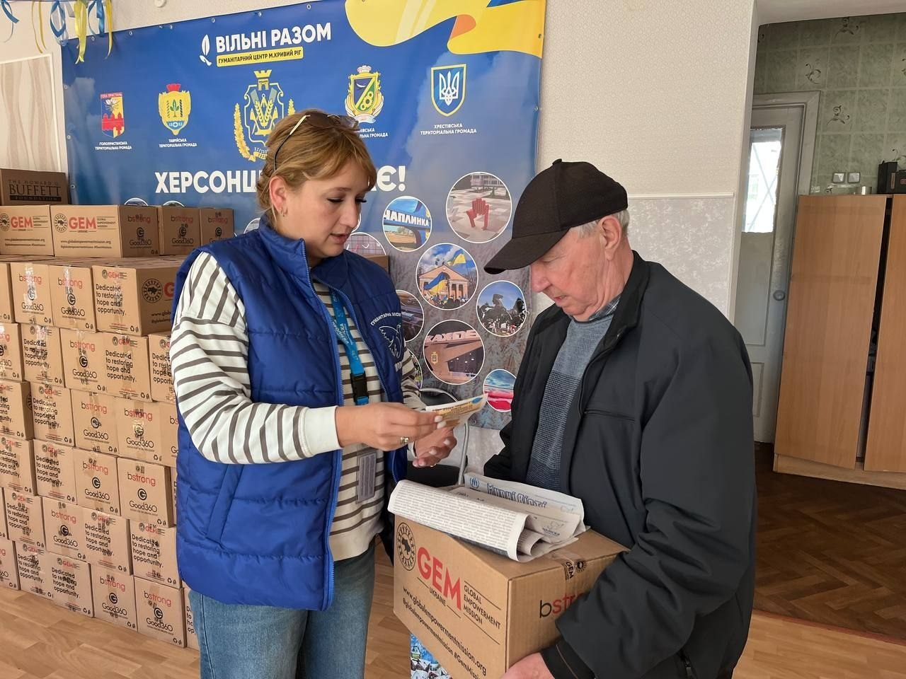 Жінка у синій волонтерській жилетці видає «Картку херсонця» літньому чоловікові, який тримає в руках коробку з гуманітарною допомогою. Дія відбувається у приміщенні на фоні інформаційного банера та стосів картонних пакунків.