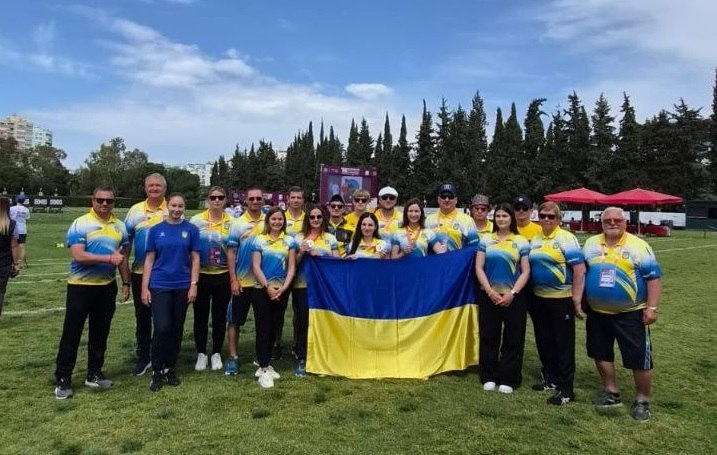 Групове фото спортивної делегації на зеленому полі під відкритим небом. Учасники, більшість з яких одягнена у синьо-жовту форму, тримають перед собою великий державний прапор України. На задньому плані видно ряд дерев, міські будівлі та спортивні намети.