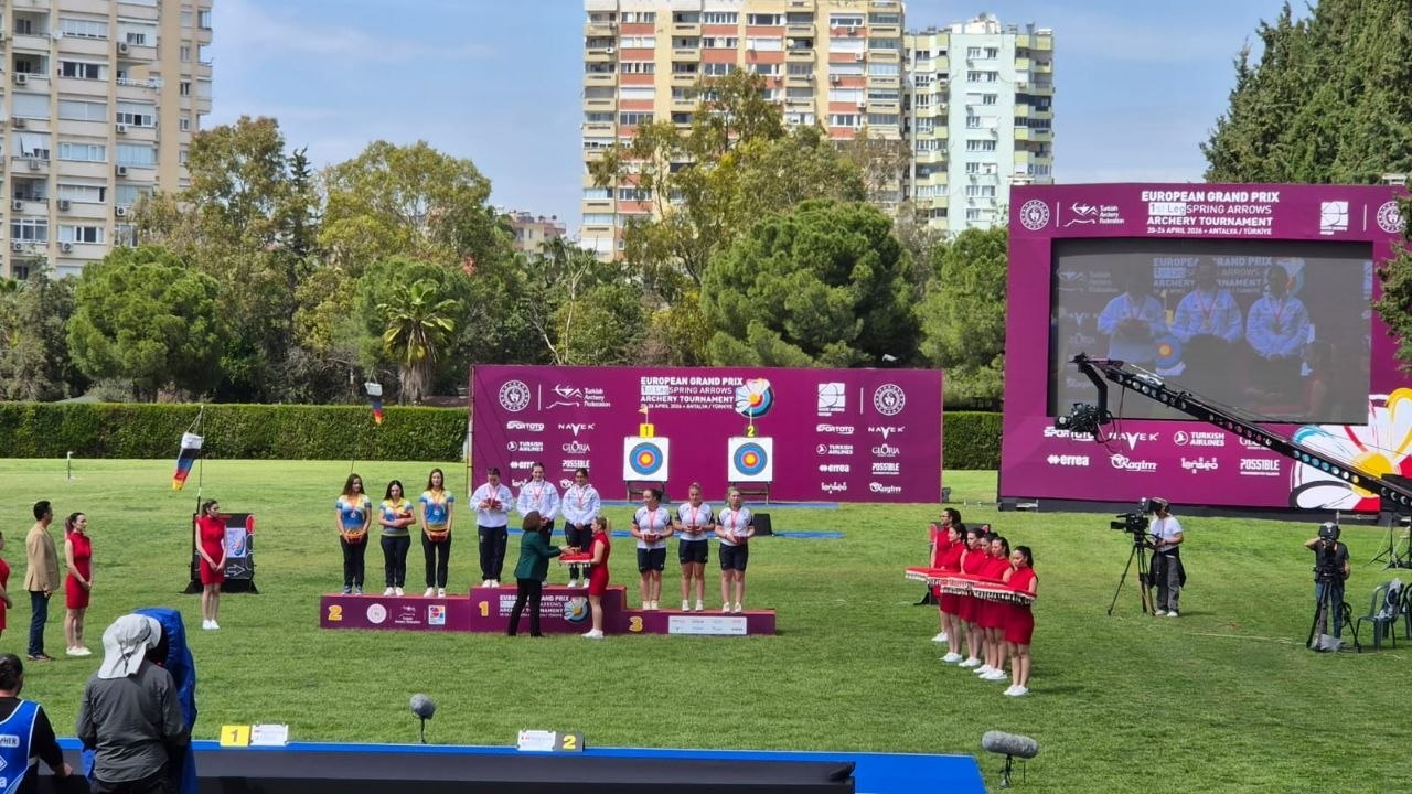 рочиста церемонія нагородження на змаганнях зі стрільби з лука. На відкритому газоні встановлено п'єдестал, де стоять три команди спортсменок. На задньому плані — великий інформаційний екран, мішені та міська забудова. Поруч знаходяться офіційні особи, асистентки в червоних сукнях та знімальна група з камерами.