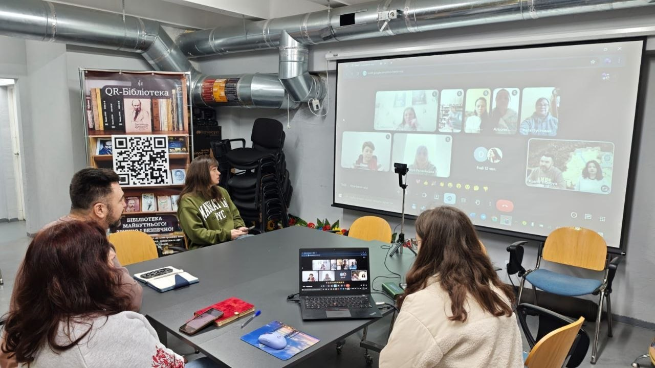 Онлайн-зустріч групи людей у сучасному приміщенні (ймовірно, укритті). Учасники за столом спілкуються через відеозв'язок, що транслюється на великий екран. Поруч розташований банер «QR-бібліотека».