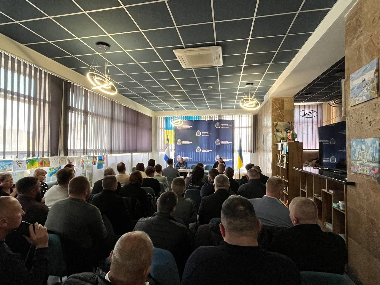 Збори великої групи людей у просторому залі з панорамними вікнами. Учасники сидять спиною до камери, дивлячись на двох спікерів за столом на фоні офіційного синього банера та державного прапора України.