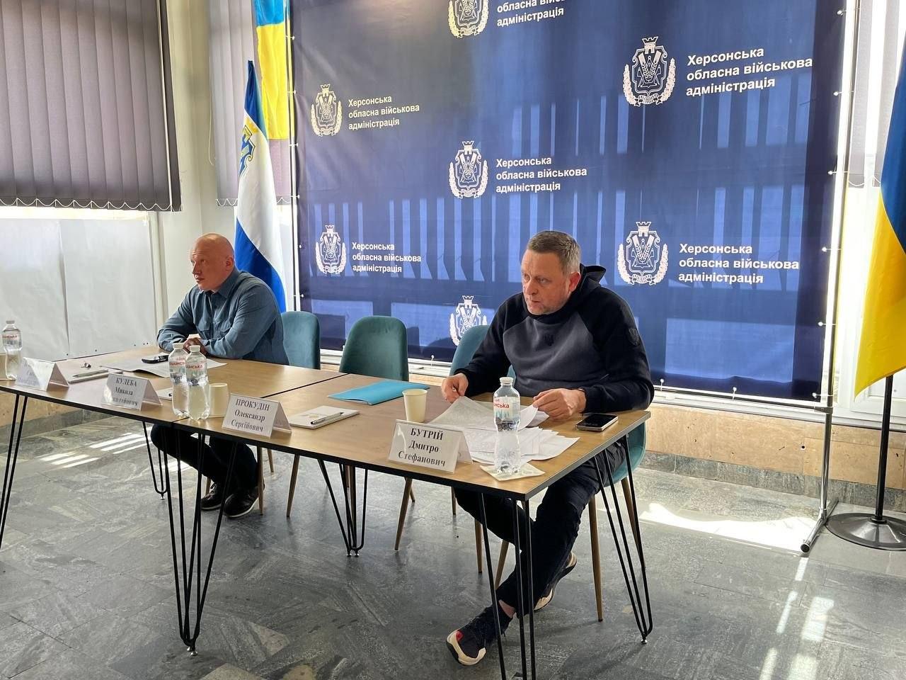 Робоча зустріч двох чоловіків за столом у офіційному приміщенні. Учасники сидять на фоні синього банера з гербами та державного прапора України. На столі перед ними лежать папери, стоять пляшки з водою та таблички з іменами.