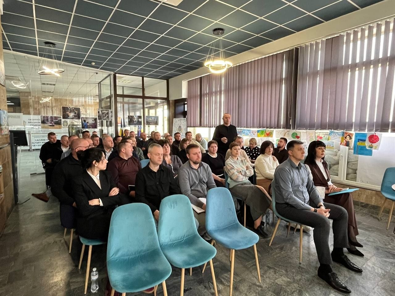 Збори групи людей у просторій залі. Учасники сидять у рядах і слухають виступ спікера. Уздовж великих вікон із вертикальними жалюзі розміщена виставка дитячих малюнків. Приміщення має сучасний інтер'єр із плитковою стелею та кільцеподібними люстрами.