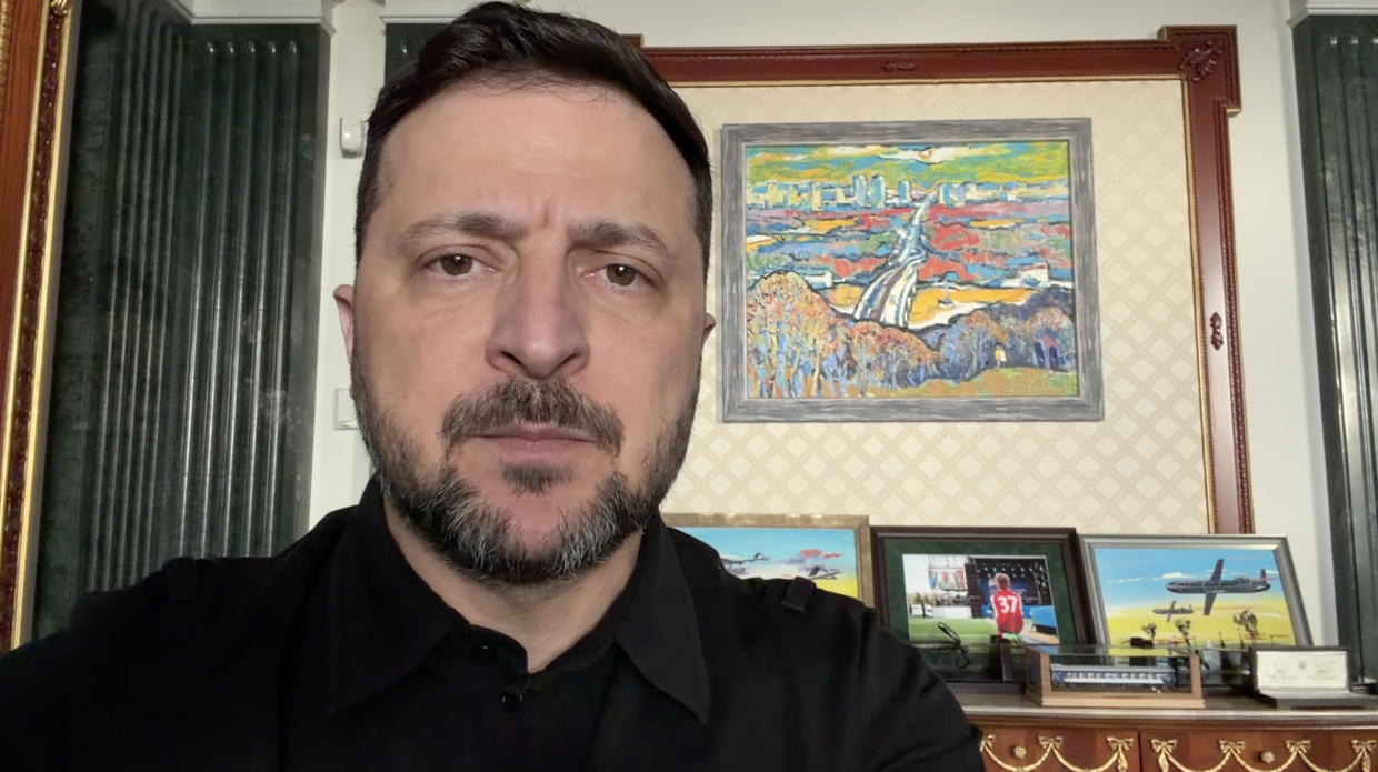 Чоловік у чорній сорочці на фоні кабінету з класичним інтер'єром. На стіні за ним висить яскрава картина з пейзажем, а на комоді під нею розставлені світлини у рамках та макети військової техніки.