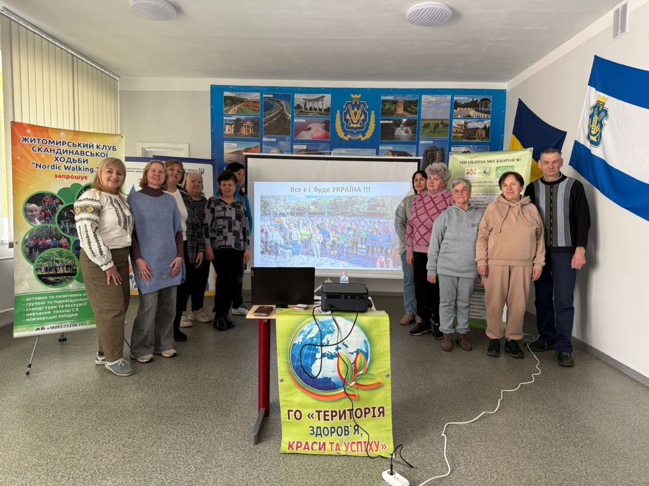 Групове фото учасників заходу в приміщенні Житомирського клубу скандинавської ходьби. У залі розміщено інформаційні банери ГО «Територія здоров'я, краси та успіху», екран із презентацією та державну символіку.