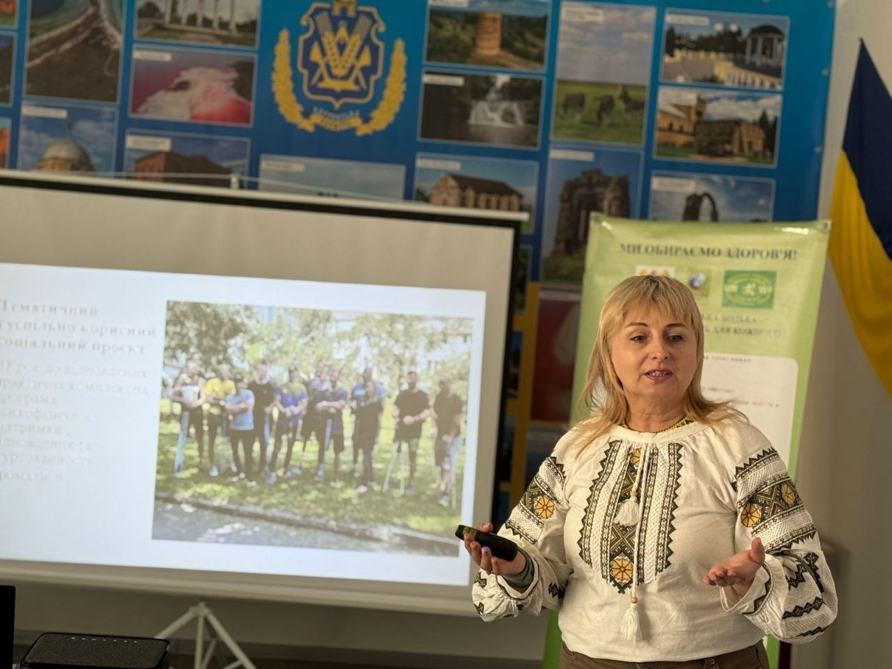 Жінка у вишиванці проводить презентацію в приміщенні Житомирського клубу скандинавської ходьби. Позаду неї — екран із тематичним слайдом, банер «Ми обираємо здоров'я!» та стенд із фотографіями й державною символікою.