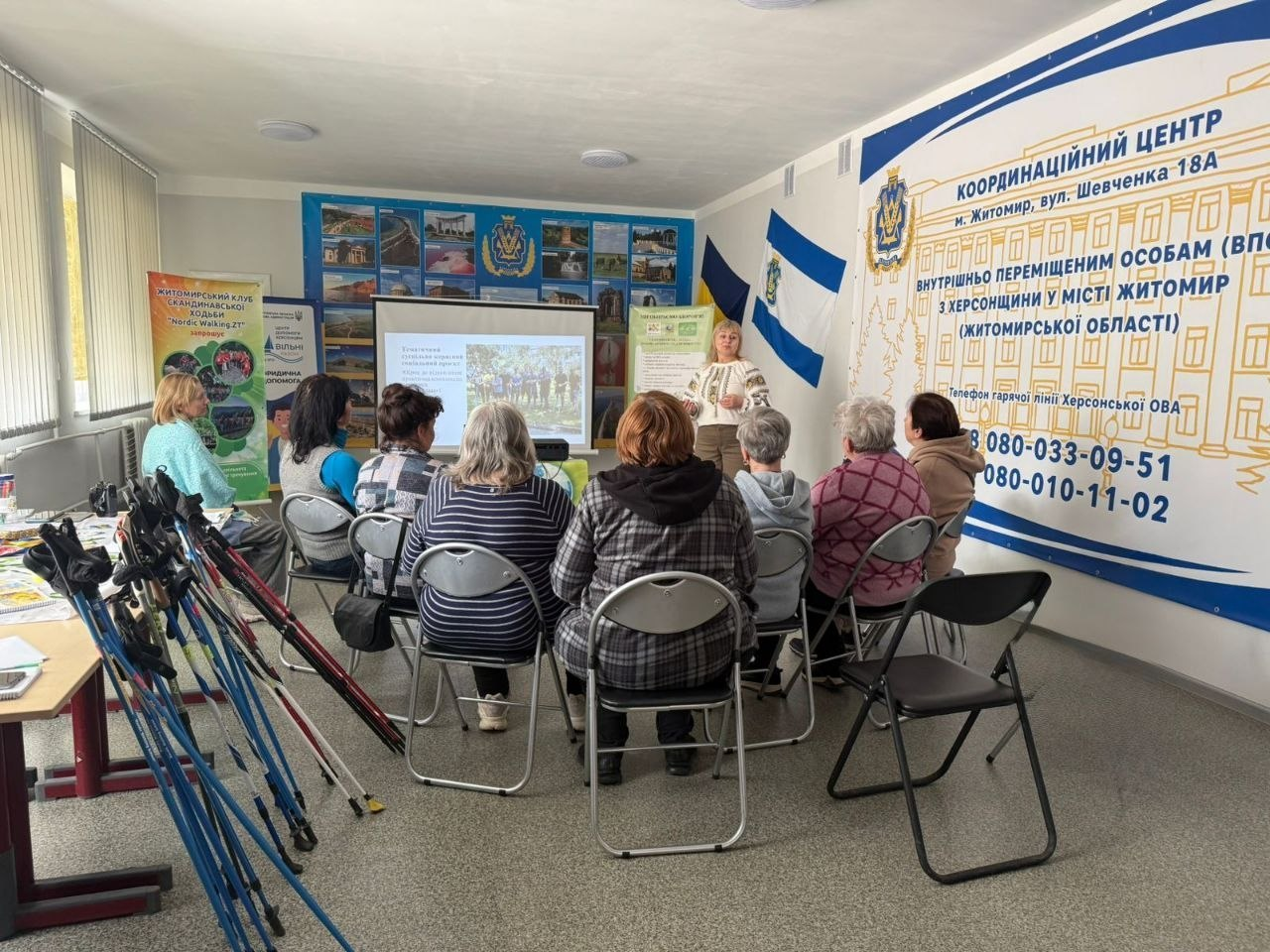 Презентація для внутрішньо переміщених осіб у Координаційному центрі м. Житомира. Слухачі сидять перед екраном, де лекторка розповідає про скандинавську ходьбу; поруч стоять палиці для занять та інформаційний банер із контактами гарячої лінії Херсонської ОВА.