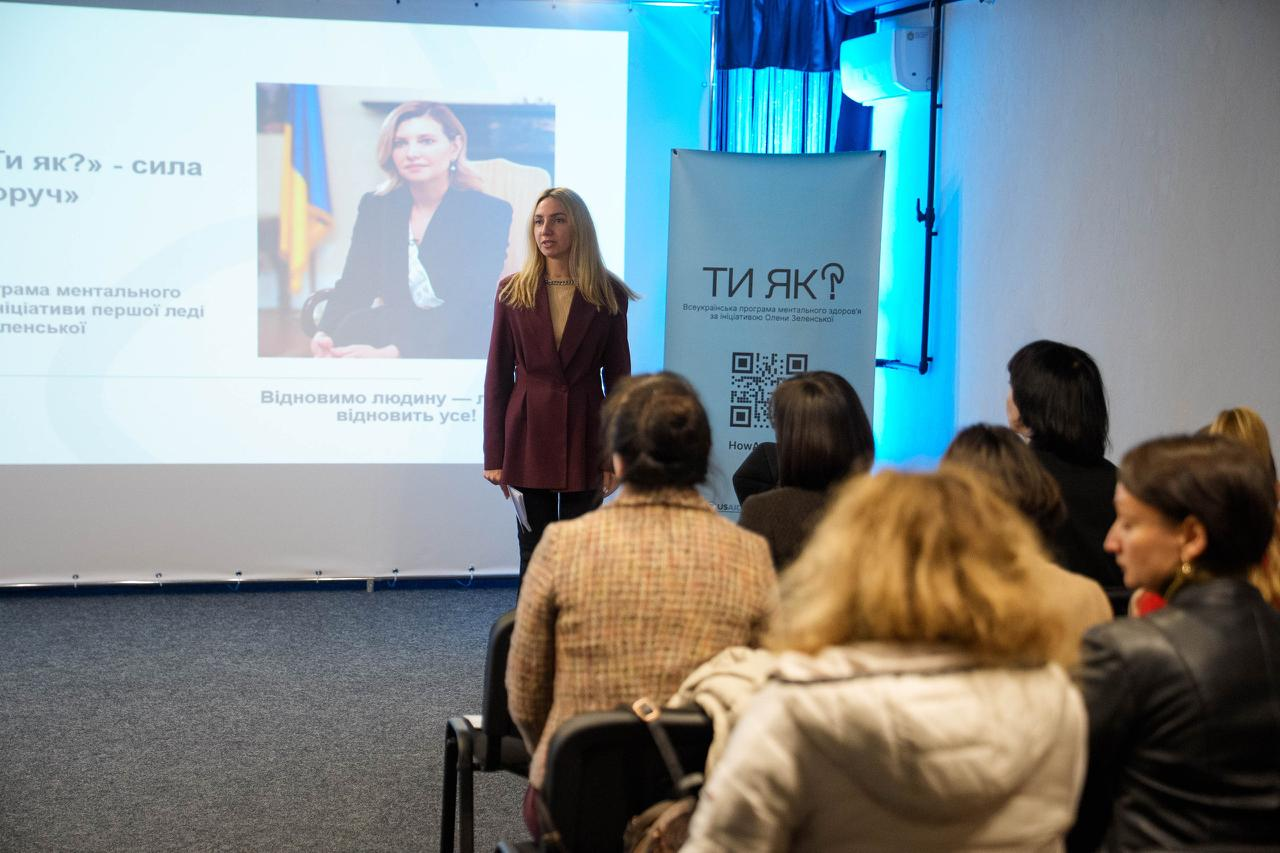 Презентація програми ментального здоров’я «Ти як?» для аудиторії. Спікерка виступає перед групою людей на фоні екрана з тематичним слайдом та банера з QR-кодом проєкту.
