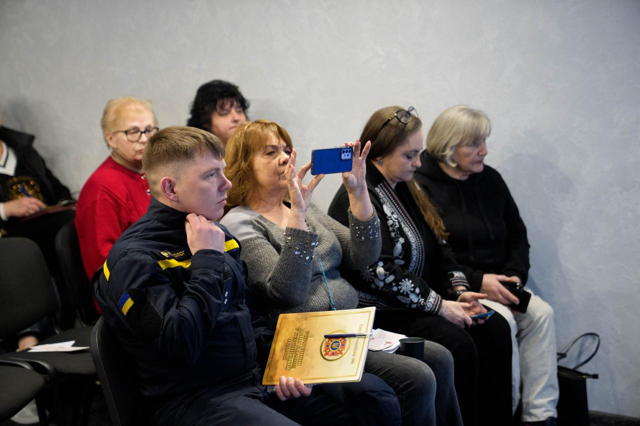 Група учасників під час офіційного заходу. На передньому плані — представник ДСНС у формі з папкою, поруч жінка фіксує презентацію на смартфон, а на задньому плані сидять інші слухачі, одна з яких у вишиванці.