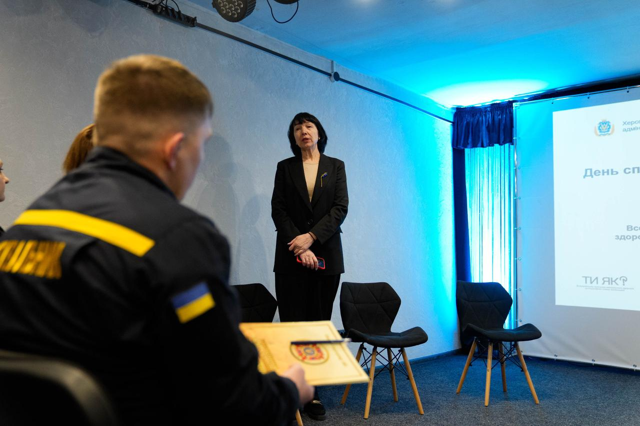 Виступ спікерки перед представниками ДСНС під час заходу, присвяченого ментальному здоров'ю. На фоні — екран із логотипами Херсонської ОВА та програми «Ти як?», на передньому плані — рятувальник у формі з шевроном-прапором України.