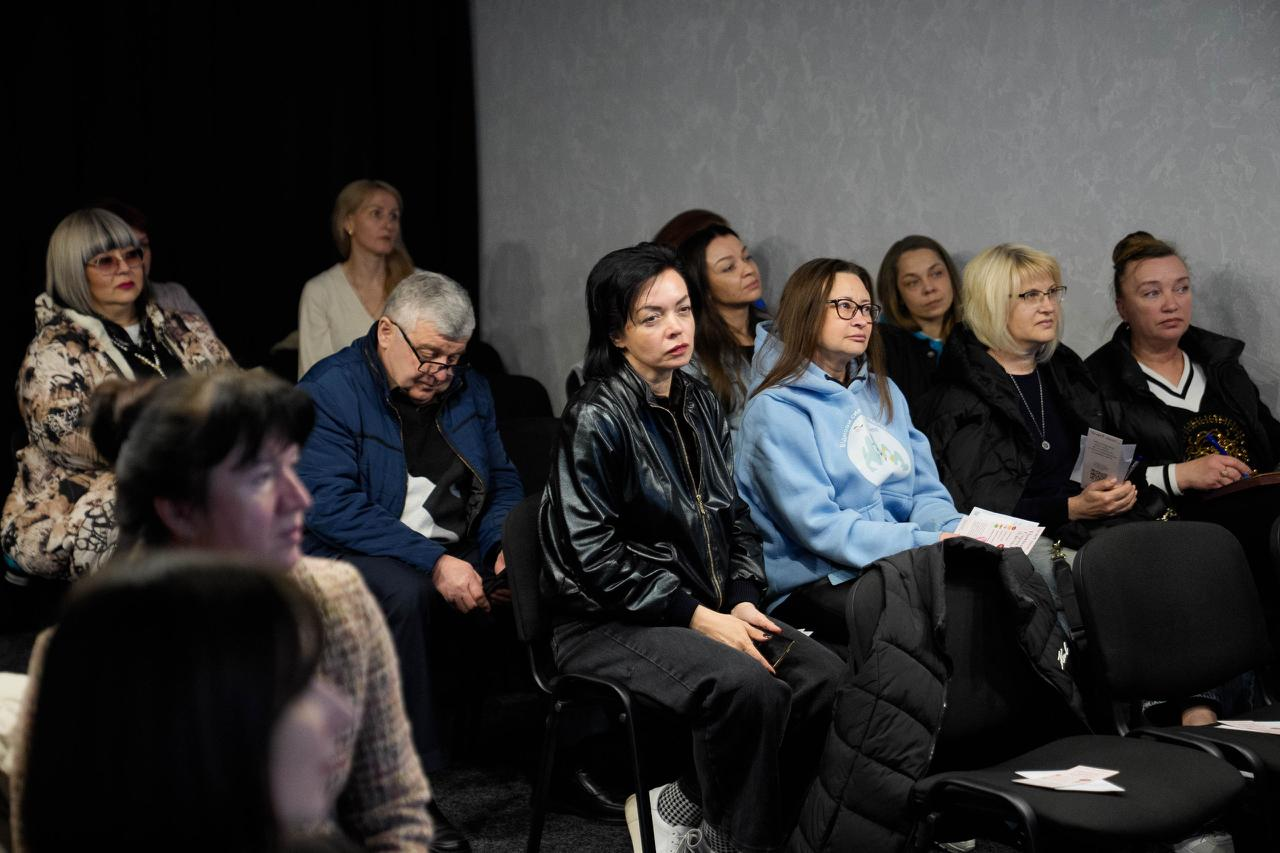 Група людей у залі уважно слухає лекцію або презентацію. Учасники заходу сидять на стільцях, дехто тримає в руках інформаційні буклети.