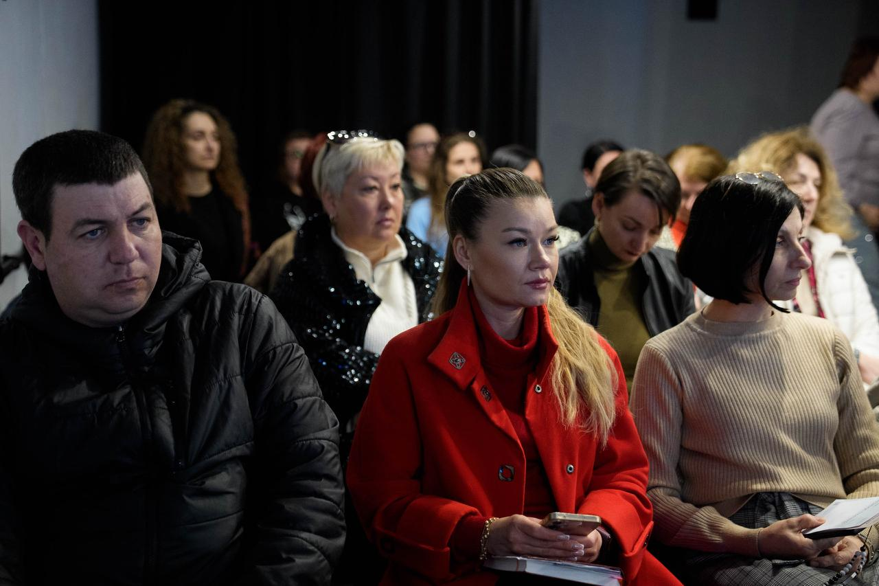 Учасники освітнього заходу або лекції в аудиторії. Люди різного віку зосереджено слухають спікера; на передньому плані — жінка в яскравому червоному пальті з блокнотом і телефоном у руках.