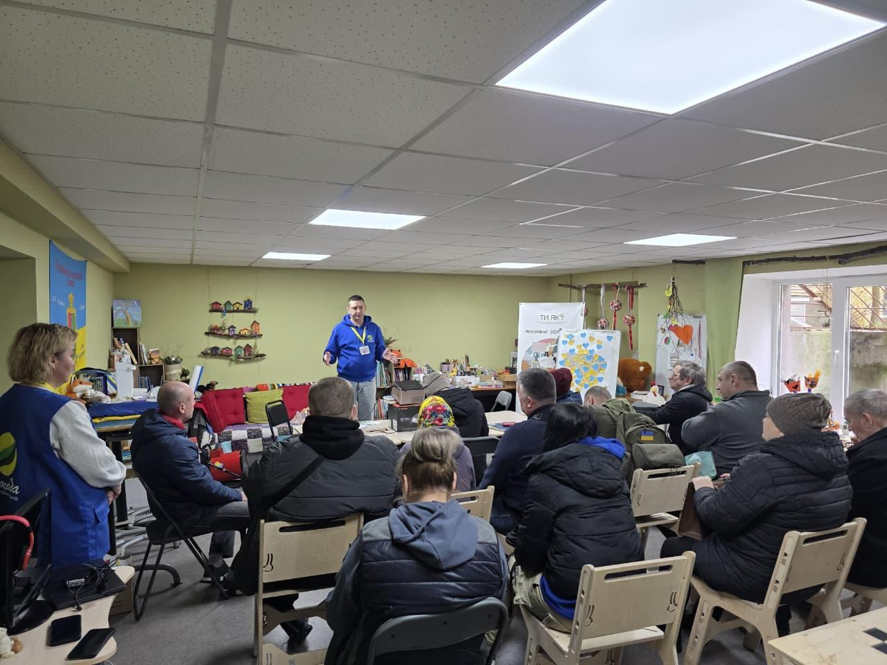 Виступ спікера перед групою слухачів у затишному приміщенні з тематичним декором. У залі розміщено стенд програми «Ти як?», дитячі вироби та патріотичну символіку.