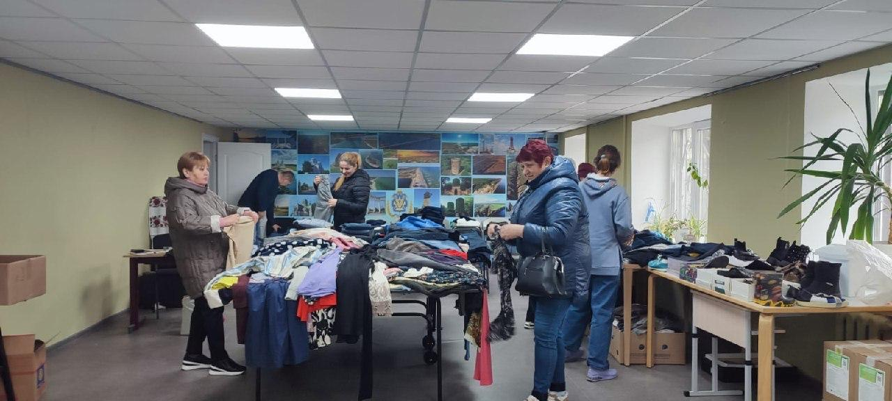 Видача гуманітарної допомоги у вигляді одягу та взуття в офісному приміщенні. Люди обирають речі, розкладені на великих столах; на задньому плані розміщена стіна з фотографіями визначних місць Херсонщини.