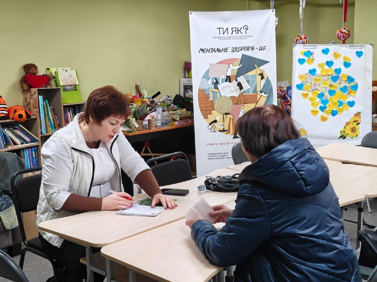 Консультування відвідувачки в межах програми ментального здоров'я «Ти як?». Фахівчиня заповнює документи за столом, поруч розташовано тематичний банер проєкту та інтерактивну карту України, прикрашену синьо-жовтими сердечками.