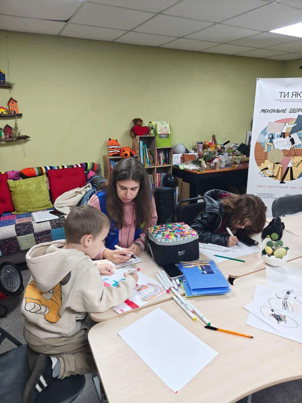 Заняття з арттерапії для дітей та дорослих у межах програми «Ти як?». Учасники малюють маркерами за столом у затишній кімнаті з книжковими полицями та яскравим декором; поруч розміщено інформаційний банер проєкту про ментальне здоров'я.