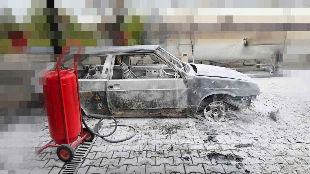 Згорілий легковий автомобіль на бруківці після пожежі. Салон повністю вигорів, колеса та шибки знищені, а кузов покритий попелом. Поруч на передньому плані стоїть великий пересувний червоний вогнегасник на колесах зі шлангом.