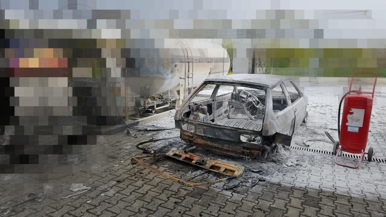 Згорілий вщент легковий автомобіль на майданчику з бруківки, вкритій білим порошком чи піною. Машина повністю знищена вогнем, без вікон та коліс. Праворуч стоїть великий червоний вогнегасник, а на задньому плані розташована велика біла цистерна.