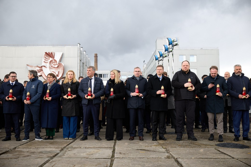Група людей у темному верхньому одязі стоїть у шеренгу на тлі промислової будівлі з декоративним барельєфом у формі птаха та освітлювальними приладами. Кожен учасник тримає в руках запалену лампадку червоного кольору.