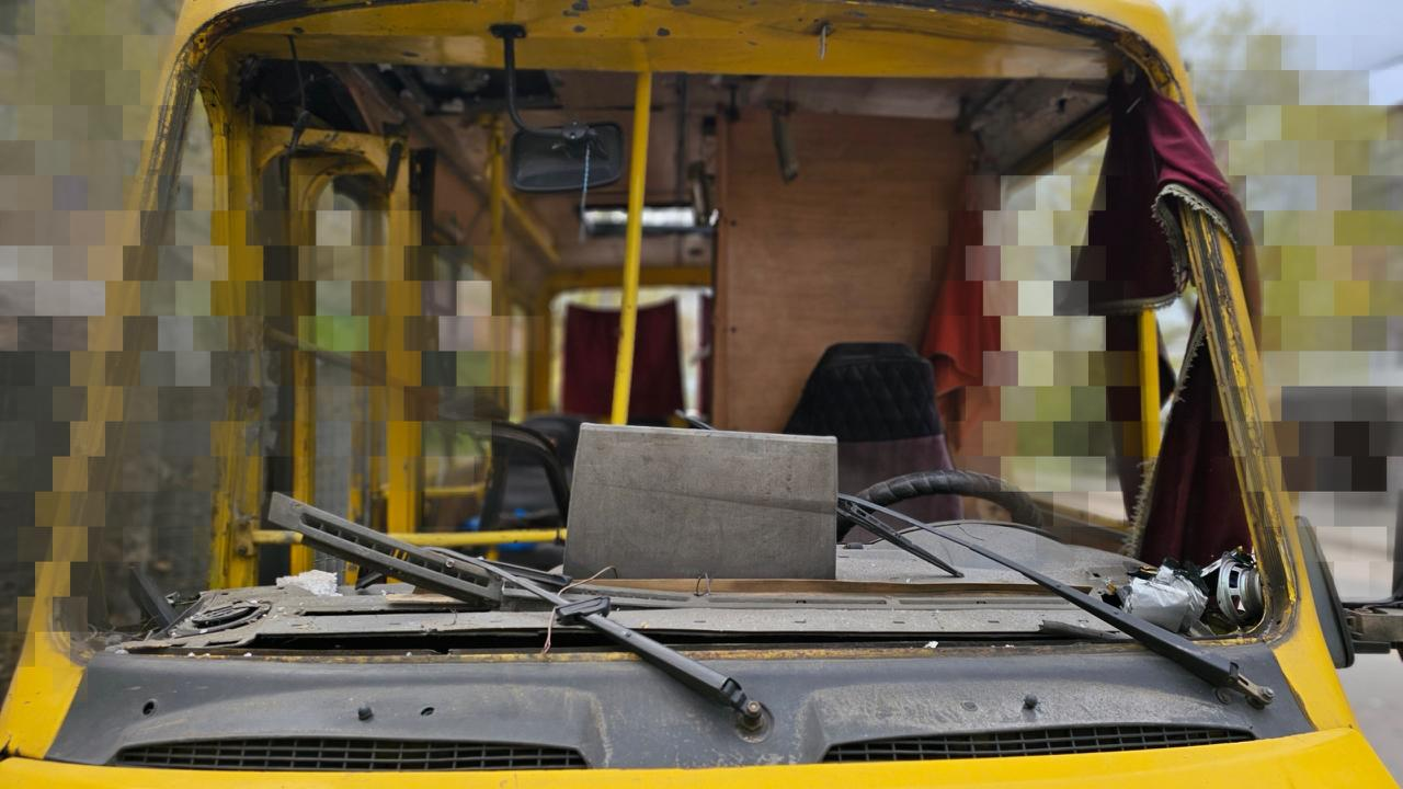 Жовтий автобус із повністю вибитим лобовим склом, через отвір якого видно пошкоджений салон. Погнуті склоочисники лежать на капоті, а краї фотографії частково приховані пікселями.