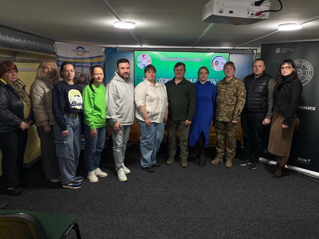 Група людей у різноманітному повсякденному та форменому одязі стоїть у шеренгу в приміщенні на тлі великого екрана з текстом «Креативний простір СИНЕРГІЯ» та двох інформаційних банерів з боків.