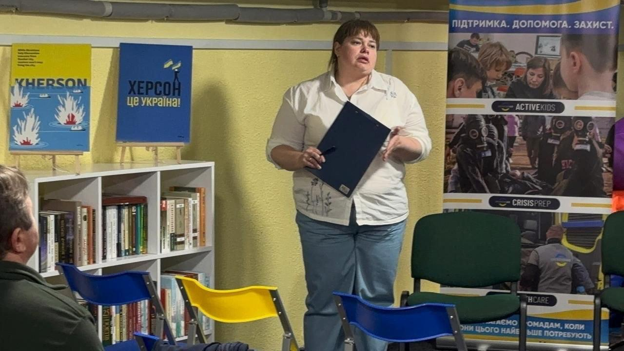 Жінка у світлій сорочці та джинсах стоїть із канцелярським планшетом у руках під час виступу в приміщенні. На тлі видно книжкові полиці, різнокольорові стільці та патріотичні плакати з написами про Херсон.