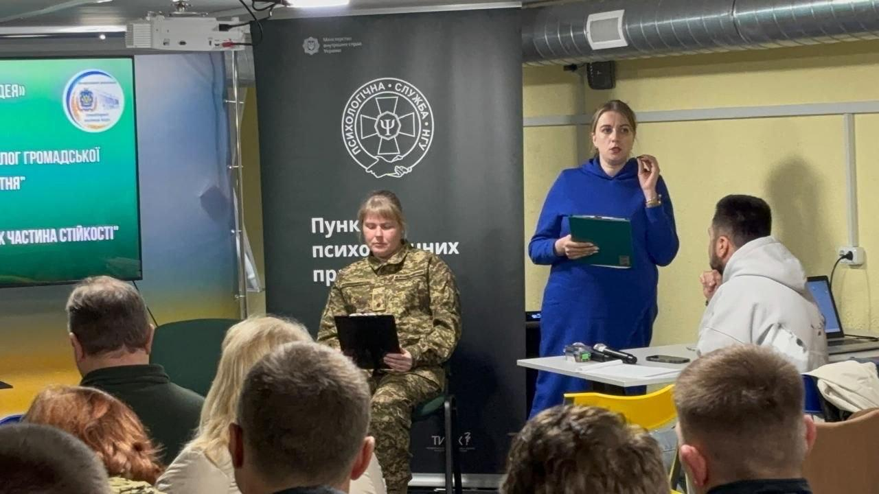 Жінка у синій сукні та жінка у військовій формі виступають перед аудиторією в приміщенні. Вони стоять і сидять на тлі великого екрана та чорного банера з емблемою Психологічної служби НГУ.