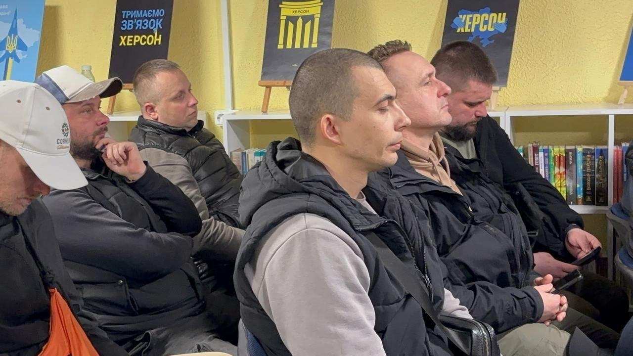 Група чоловіків у верхньому повсякденному одязі сидить у ряд під час заходу в приміщенні. На задньому плані видно полиці з книгами та патріотичні плакати з написами про Херсон.