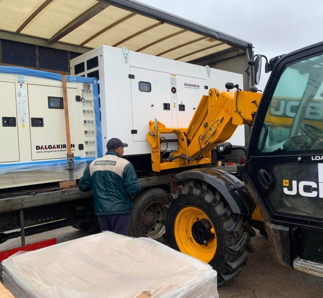 Жовтий навантажувач JCB завантажує великий білий промисловий генератор «DALGAKIRAN» у кузов вантажівки.