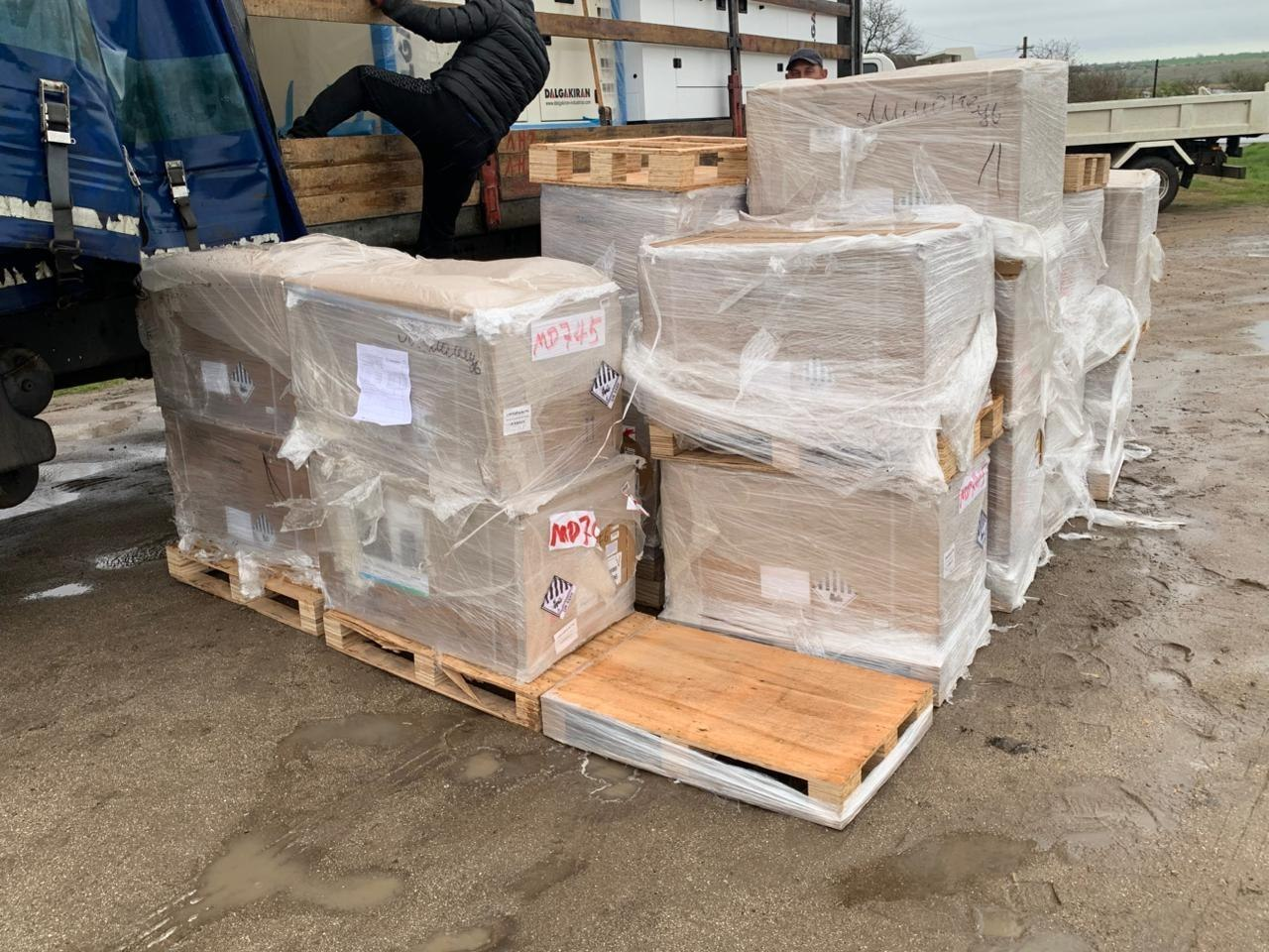 Великі стопки картонних коробок на дерев'яних піддонах, обмотані захисною плівкою, стоять просто неба біля синього вантажного автомобіля.